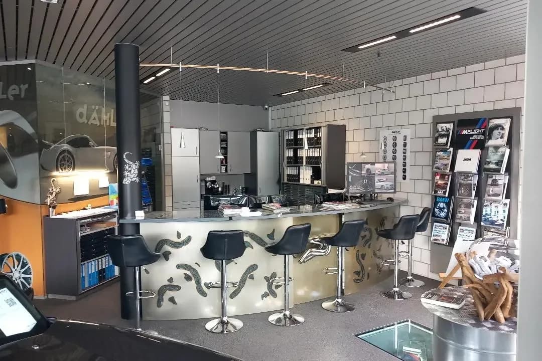 Der Empfang vor dem Umbau: weiss geflieste Wände, eine geschwungene Theke aus gebürstetem Metall mit Dekormuster, fünf schwarze Barhocker, Prospektständer und Aktenordner im Hintergrund.